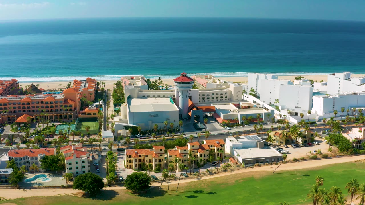 A later aerial view of Los Cabos centers white hotel-like blocks, orange-roofed structures, beach sand, and layered blue ocean, with no visible people.