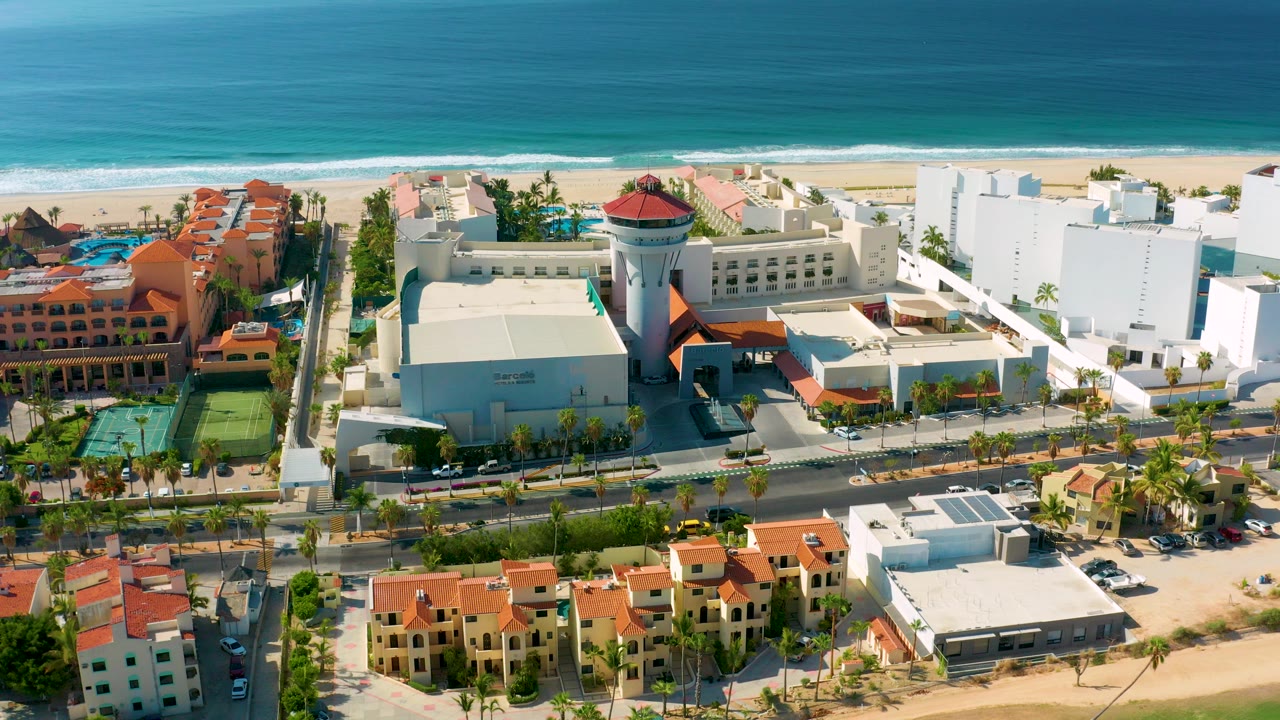 Aerial frame over Los Cabos resort buildings with terracotta roofs, tennis courts, a sandy beach, turquoise ocean, and no visible people.