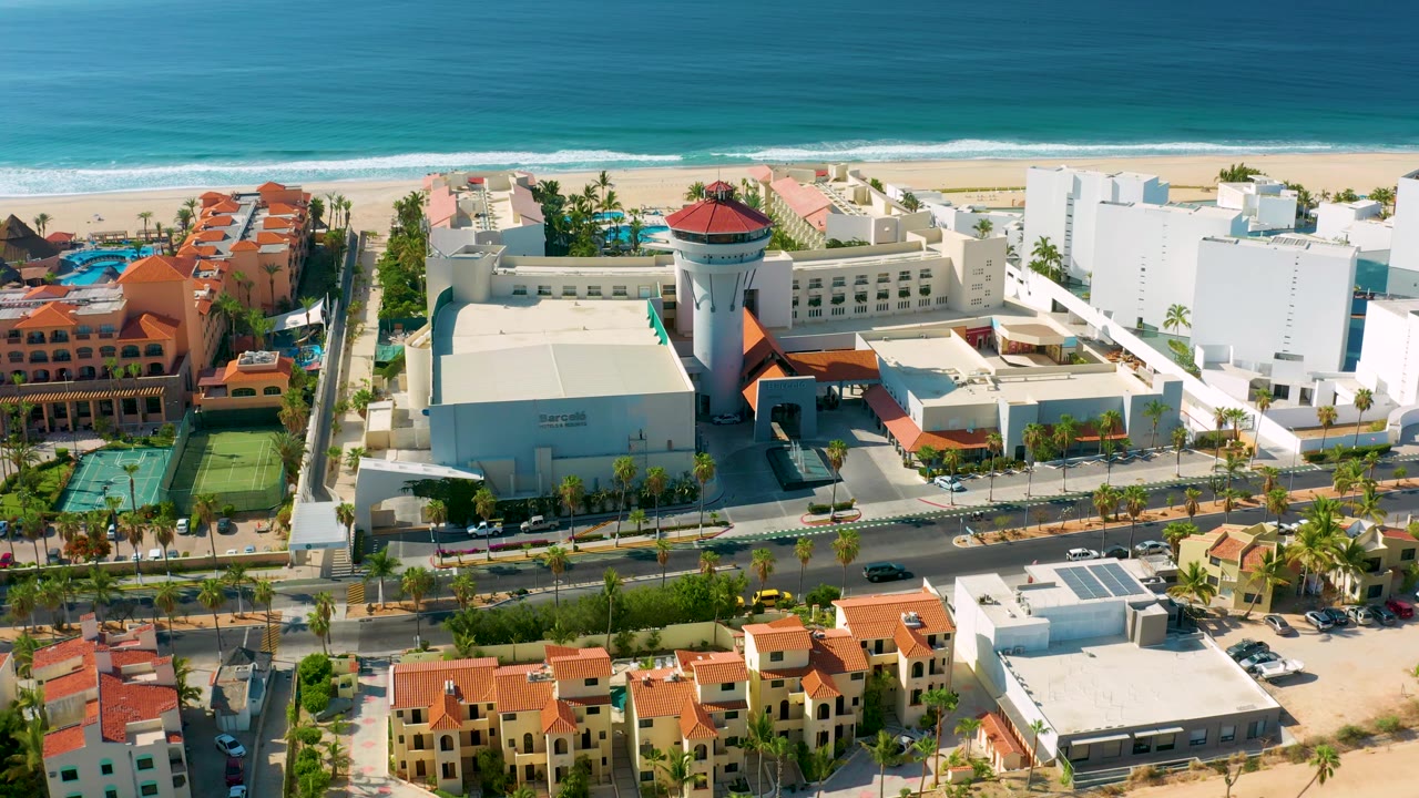 High drone frame in Los Cabos showing a resort complex, road lanes, small parked cars, beach sand, blue-green water, and no visible people.