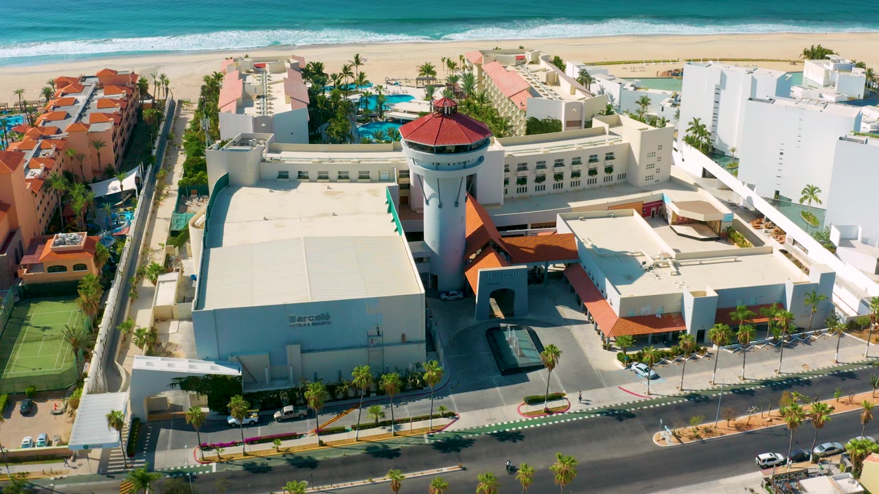 Aerial view in Los Cabos, Mexico showing a beachfront hotel complex, red-roofed round tower, pools, road, beach, turquoise ocean, and no visible people.