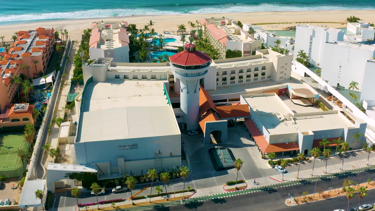 High aerial frame in Los Cabos, Mexico showing the resort tower shifted right of center, pools behind the main building, a road in front, the beach beyond, and no visible people.