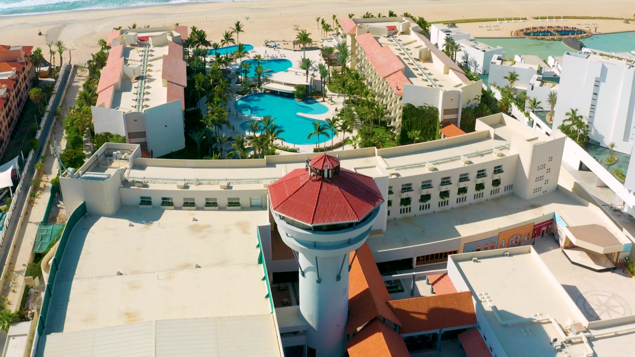 Aerial view over a Los Cabos resort with a red-roofed round tower in the foreground, white buildings, blue pools, beach, ocean surf, and tiny visible people.