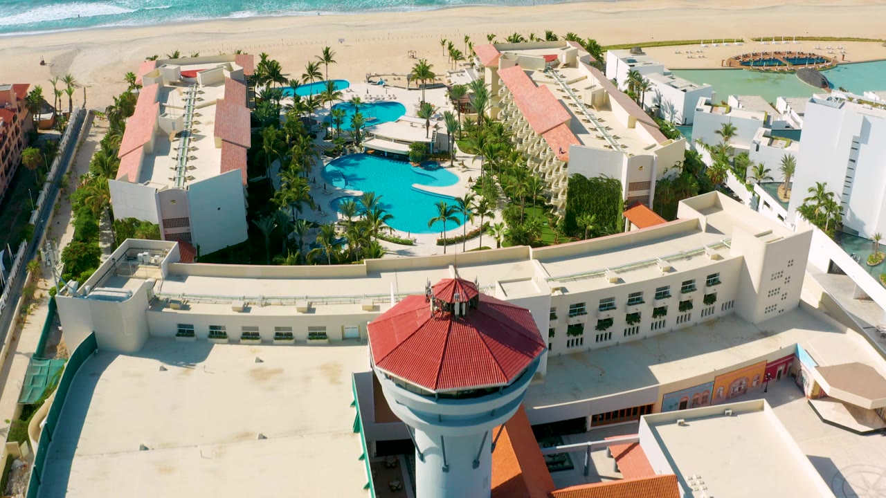 Aerial Los Cabos frame with the red-roofed tower near the lower right, white resort rooftops, curved pool areas, palm rows, beach, surf, and tiny visible guests.