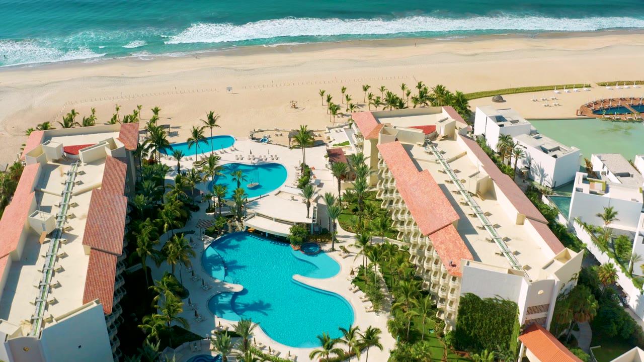 Aerial view over a Los Cabos resort with two turquoise pools, palm trees, red tile roofs, pale beach, blue-green surf, and no visible people.