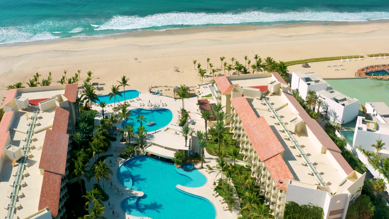 Aerial frame in Los Cabos showing red rooflines on both sides, a bright pool complex below, palm landscaping, a wide sandy beach, and no visible people.