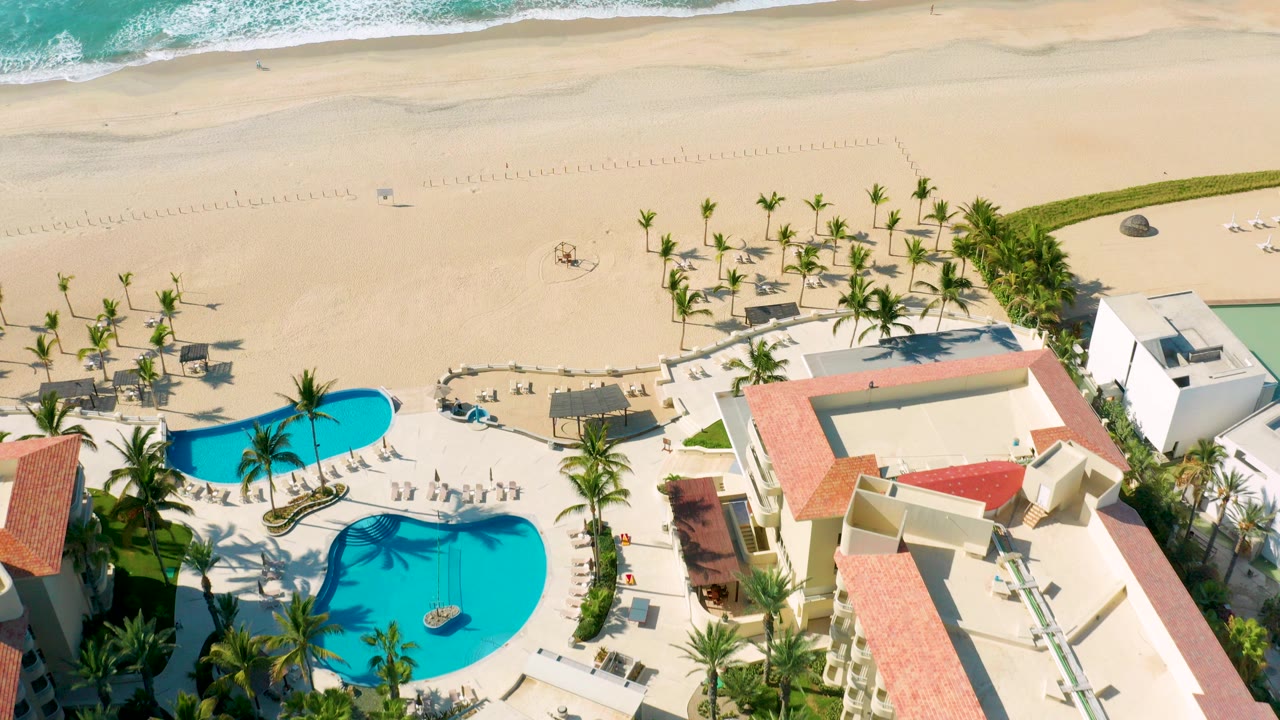 Aerial view of resort pools beside a sandy beach and turquoise surf.