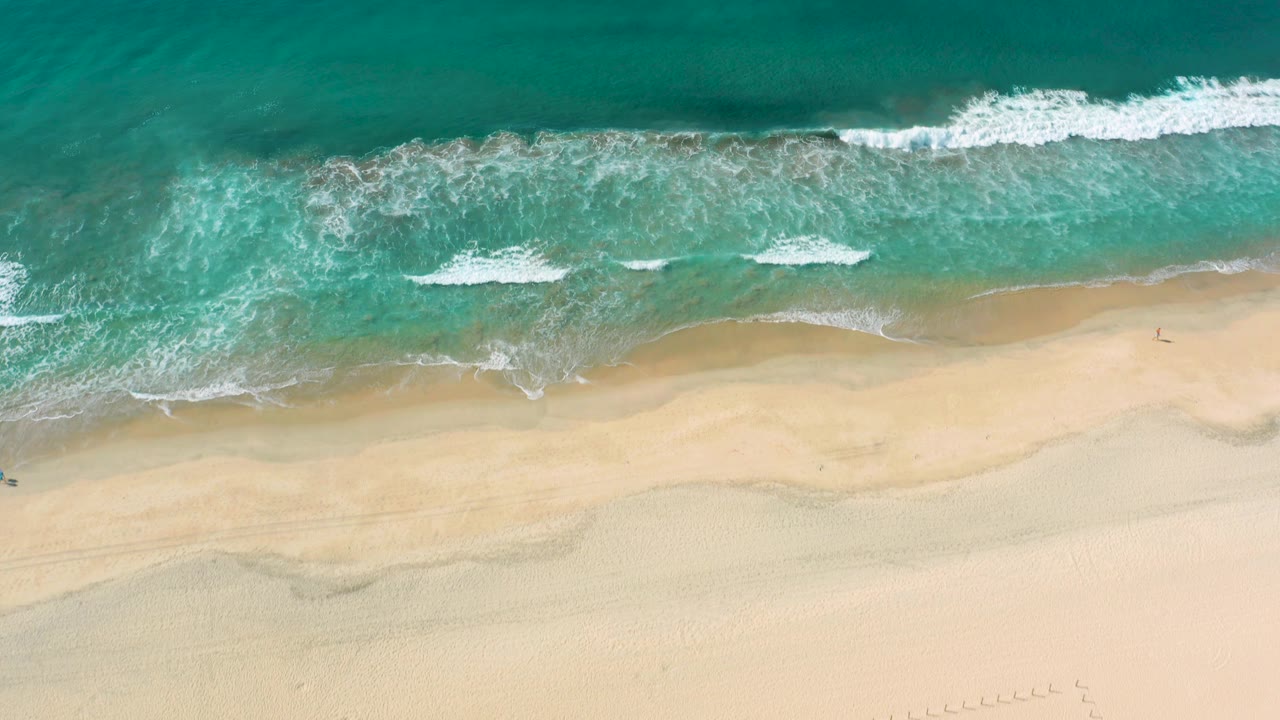 Overhead Los Cabos coast frame with teal water across the top, broken foam lines, and a wide light sand area below with no visible people.