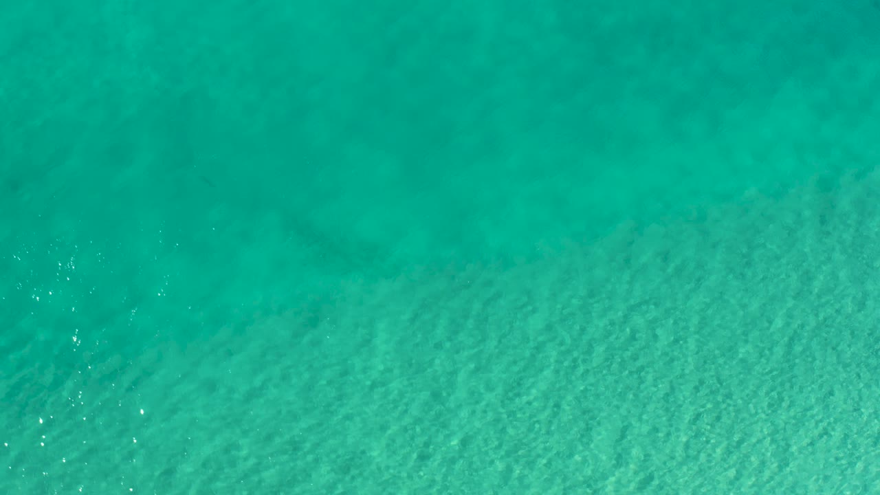Overhead aerial frame of shallow turquoise sea in Los Cabos, Mexico, with small white sun glints near the lower left and no visible people.