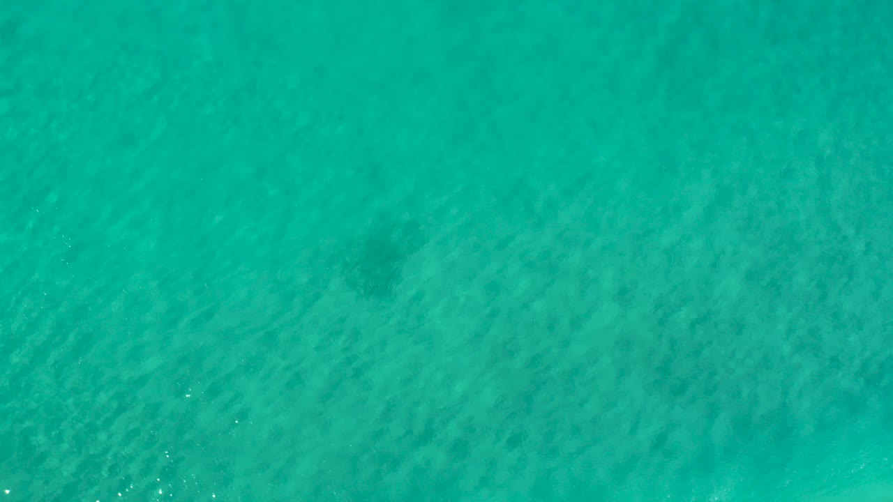 Top-down view of blue teal ocean water in Los Cabos, Mexico, with faint darker patches beneath the surface and no visible people.
