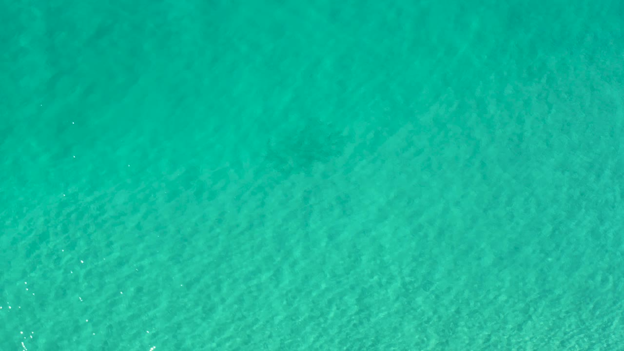 Shallow turquoise water in Los Cabos, Mexico, shows faint algae or seabed texture beneath small surface waves, no visible people.