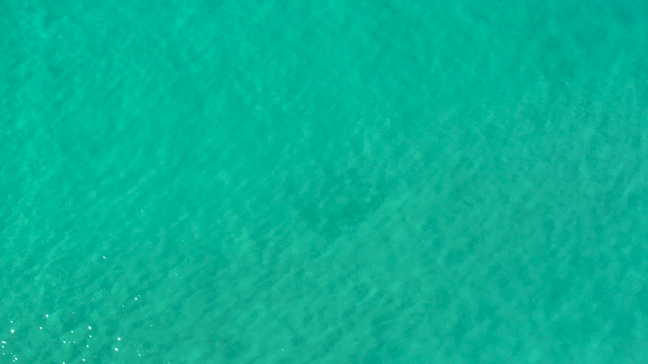 Overhead view of teal-green seawater in Los Cabos, Mexico, with faint underwater texture, small bright glints near the lower left, and no visible people.