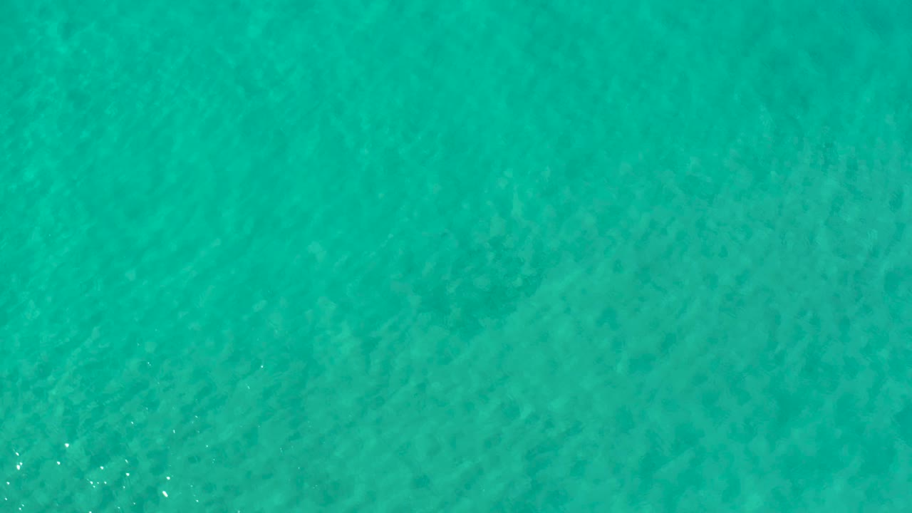 A straight-down aerial frame of clear green-blue sea in Los Cabos, Mexico, showing soft ripple patterns and darker patches beneath the surface, with no visible people.