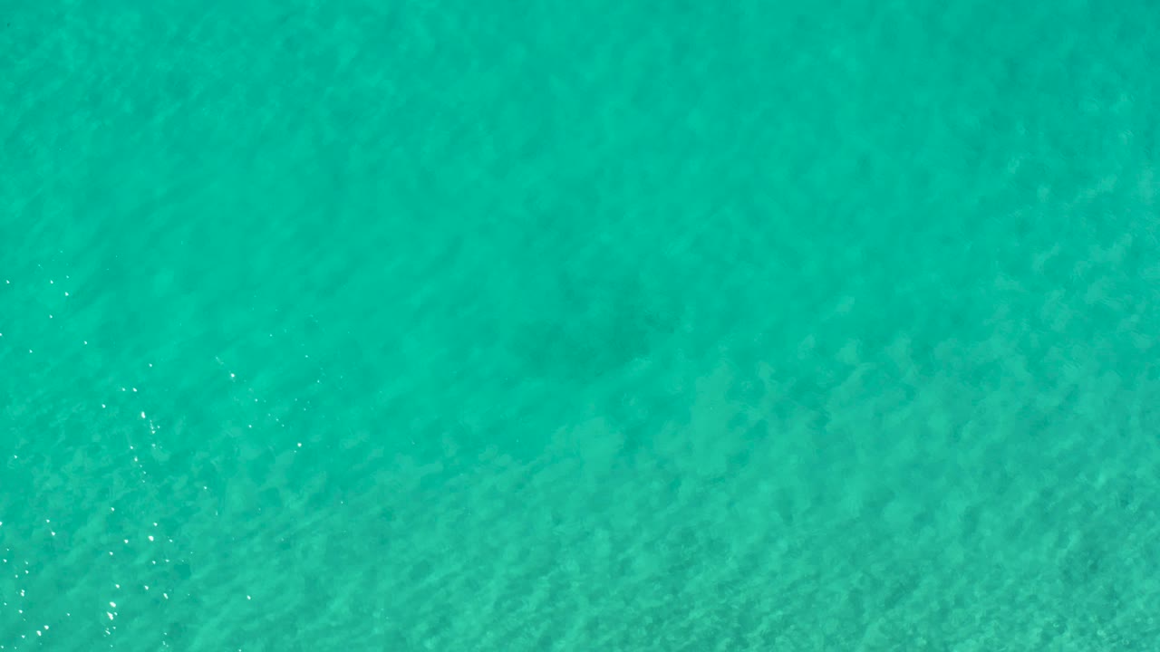 Overhead teal water in Los Cabos with gentle ripple lines, small bright flecks, and no visible shoreline or people.