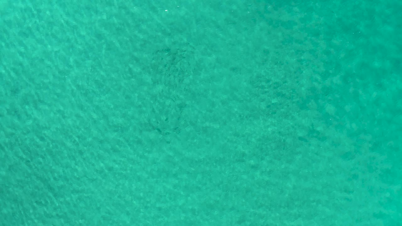 Aerial view over green-blue ocean in Los Cabos, Mexico, with mottled darker shapes visible beneath the water and no visible people.