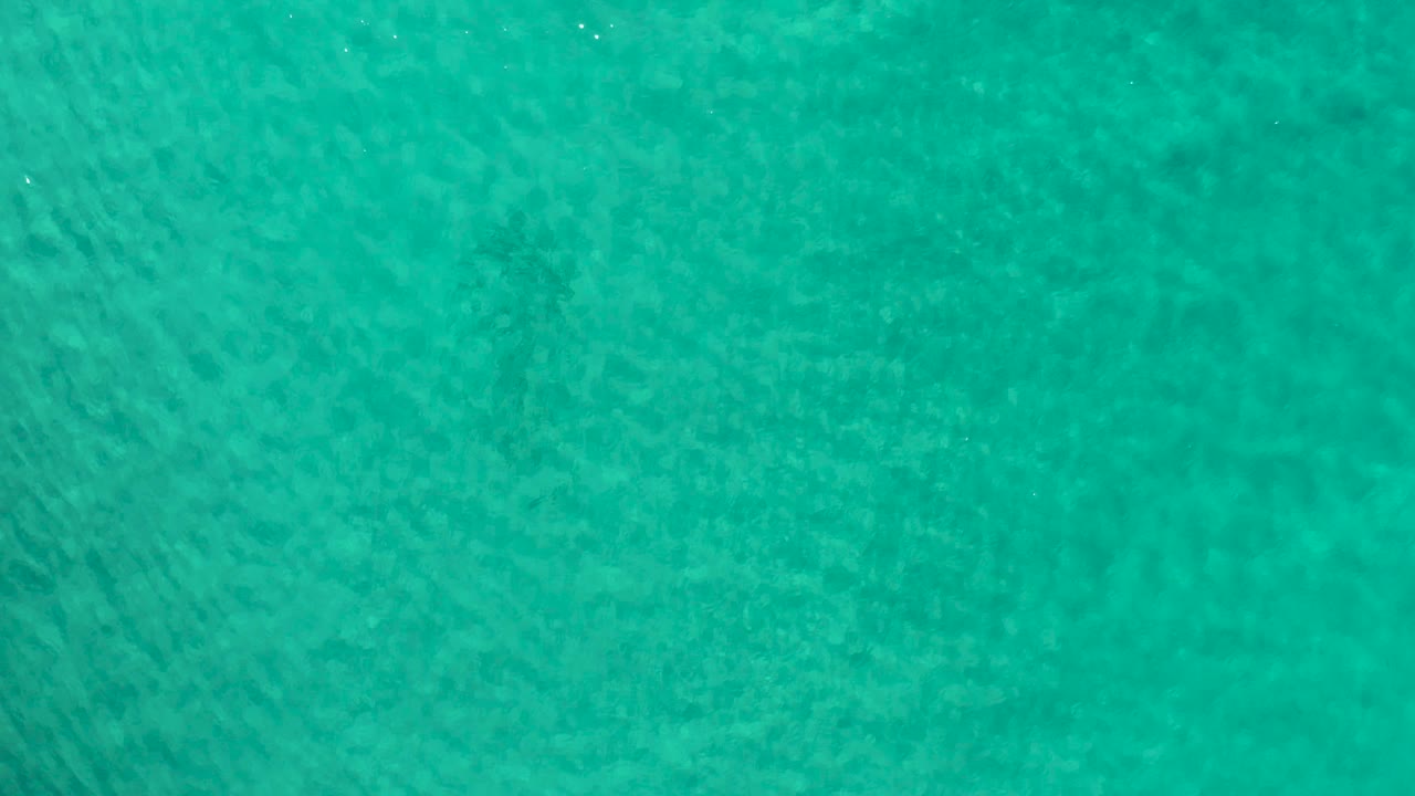 Top-down Los Cabos ocean view with turquoise water, darker underwater patches near the center, and no visible people.