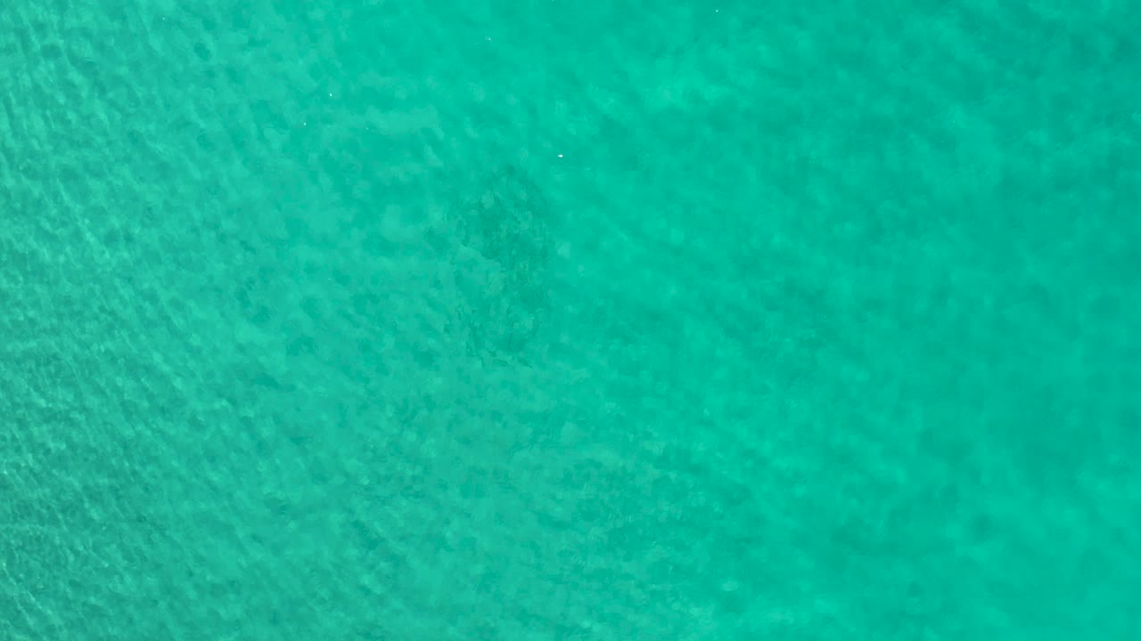 Top-down Los Cabos water view with a wider field of turquoise sea, faint mottled texture below the surface, and no visible people.