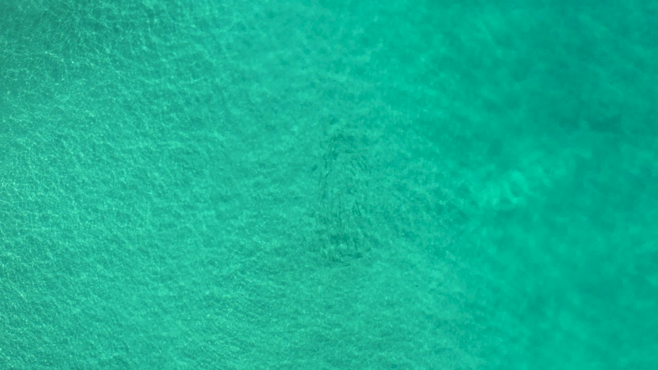Top-down view of turquoise coastal water in Los Cabos, Mexico, with faint seabed patches and no visible people.