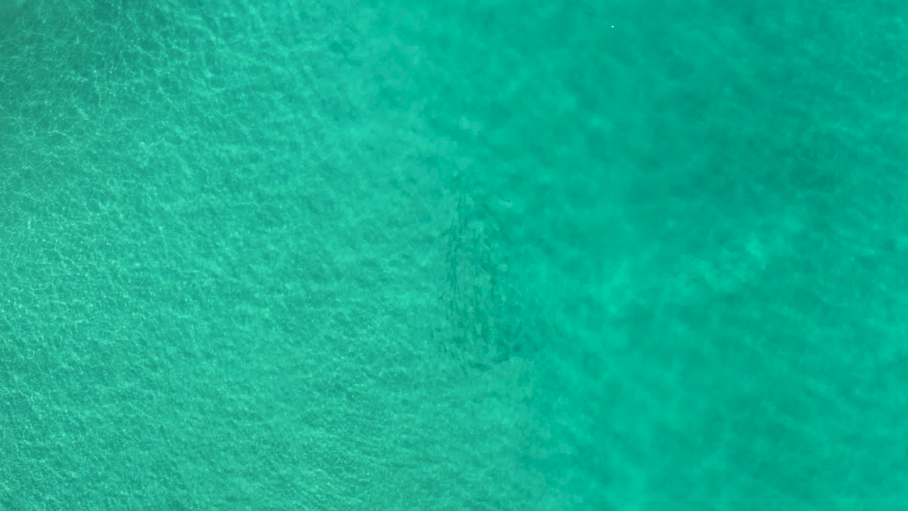 Clear turquoise water in Los Cabos, Mexico, shows darker underwater patches beneath a lightly rippled surface, with no visible people.