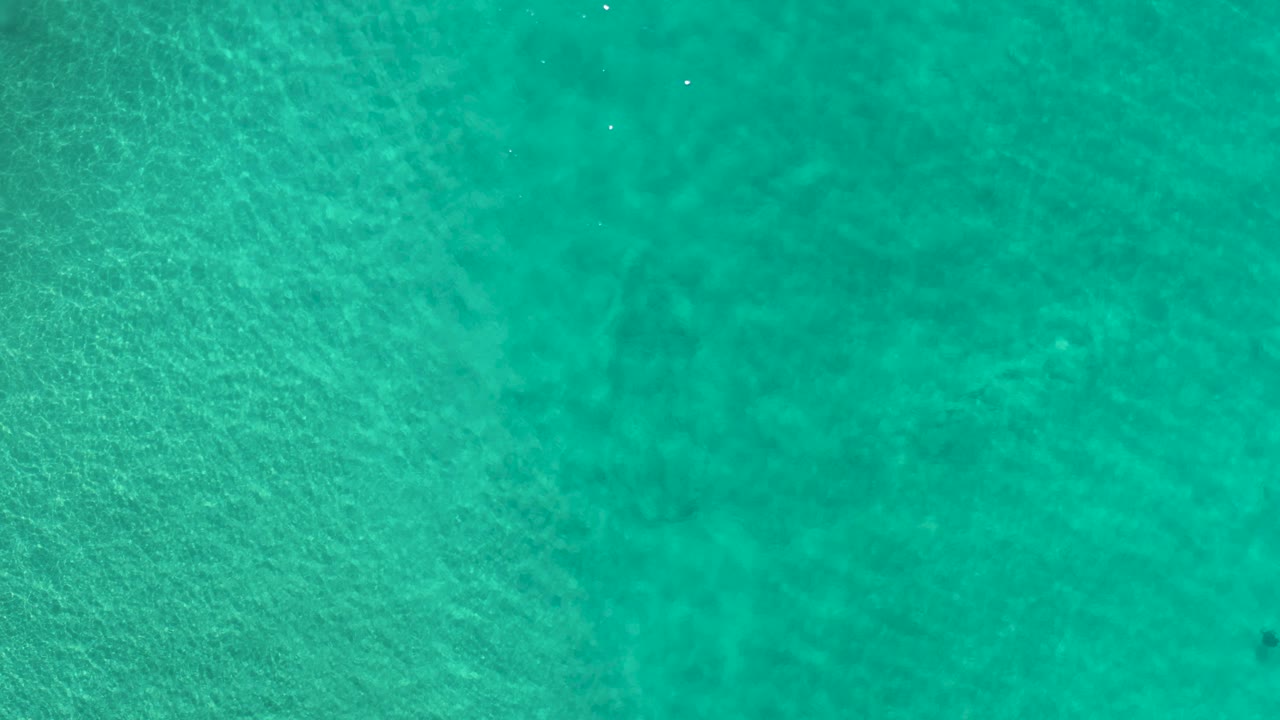 Aerial top-down frame of shallow teal water in Los Cabos, Mexico, with a denser band of seabed texture and no visible people.
