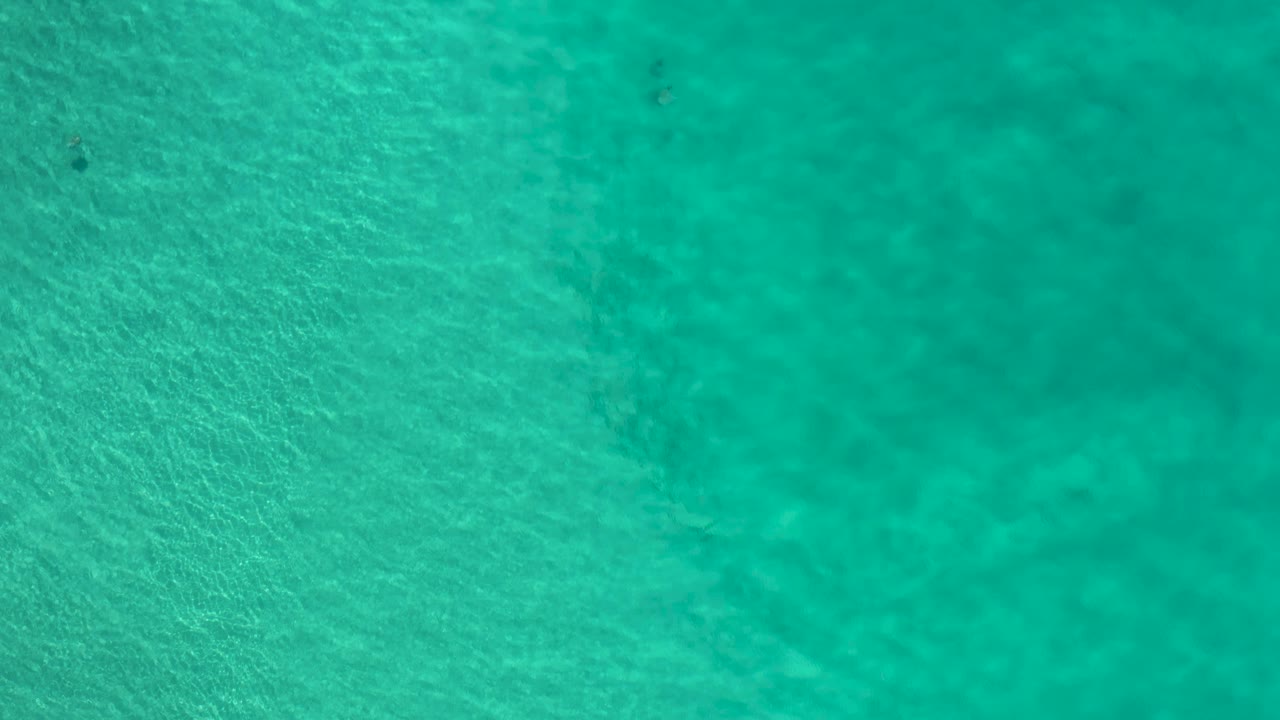 Green turquoise water seen from above in Los Cabos, Mexico, with subtle ripples, faint seabed detail, a tiny dark fish-like shape, and no visible people.