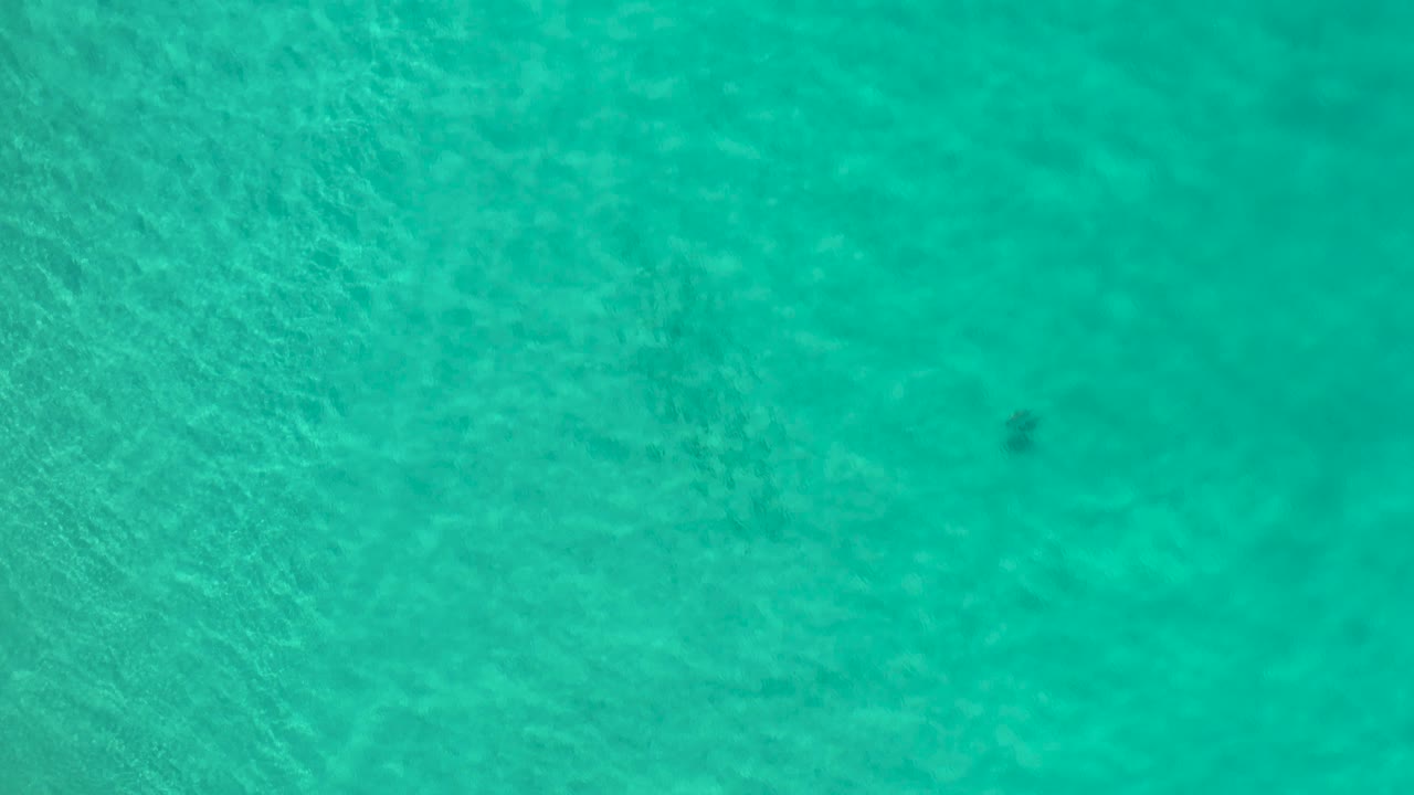 Aerial view of shallow turquoise water in Los Cabos with diffuse bottom detail and a small dark patch near the top right, no visible people.