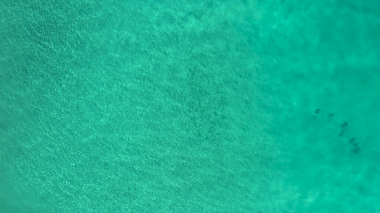 Overhead view of clear turquoise sea water in Los Cabos, Mexico, with small people visible as dark shapes near the upper center of the frame.