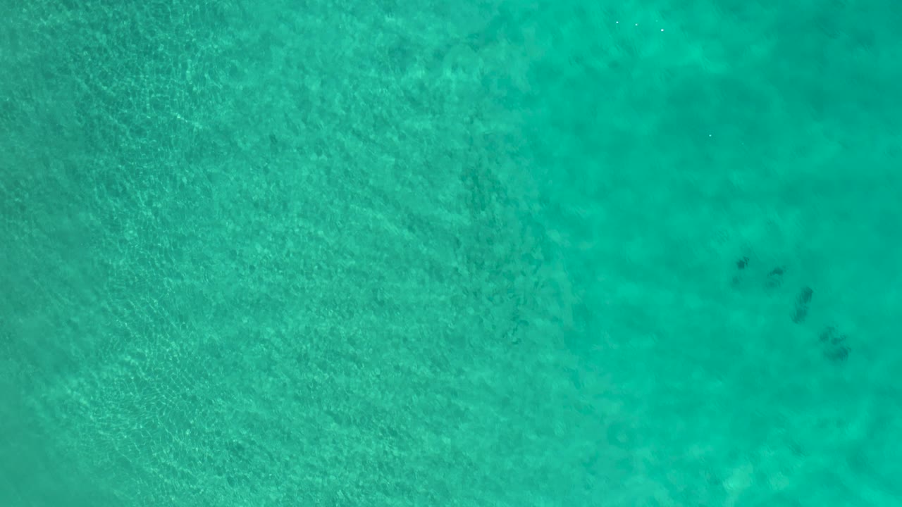 High overhead view of turquoise sea water in Los Cabos, Mexico, with tiny people visible near the top-right edge and darker patches below the surface.