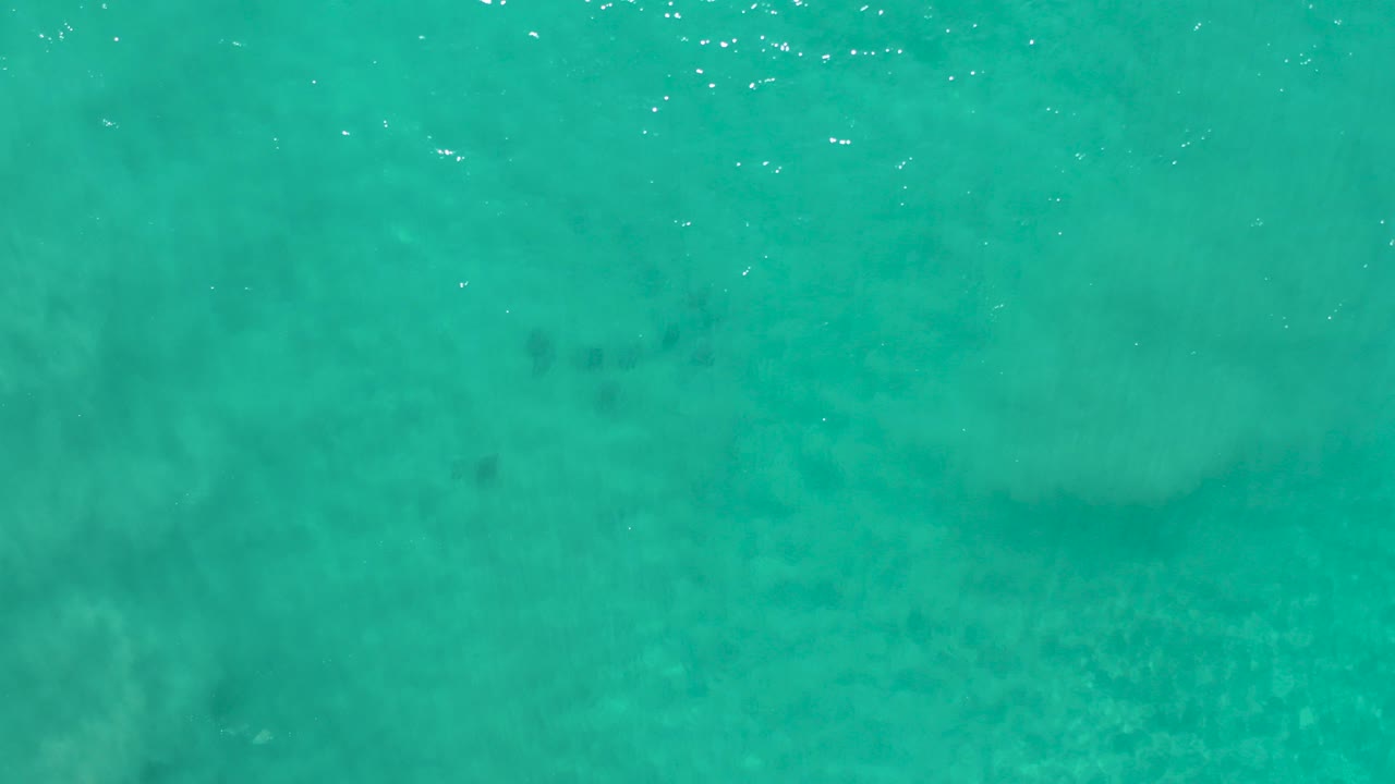 Top-down view of turquoise water in Los Cabos, Mexico, with pale sandy patches below the surface, small white ripples near the top edge, and no visible people.