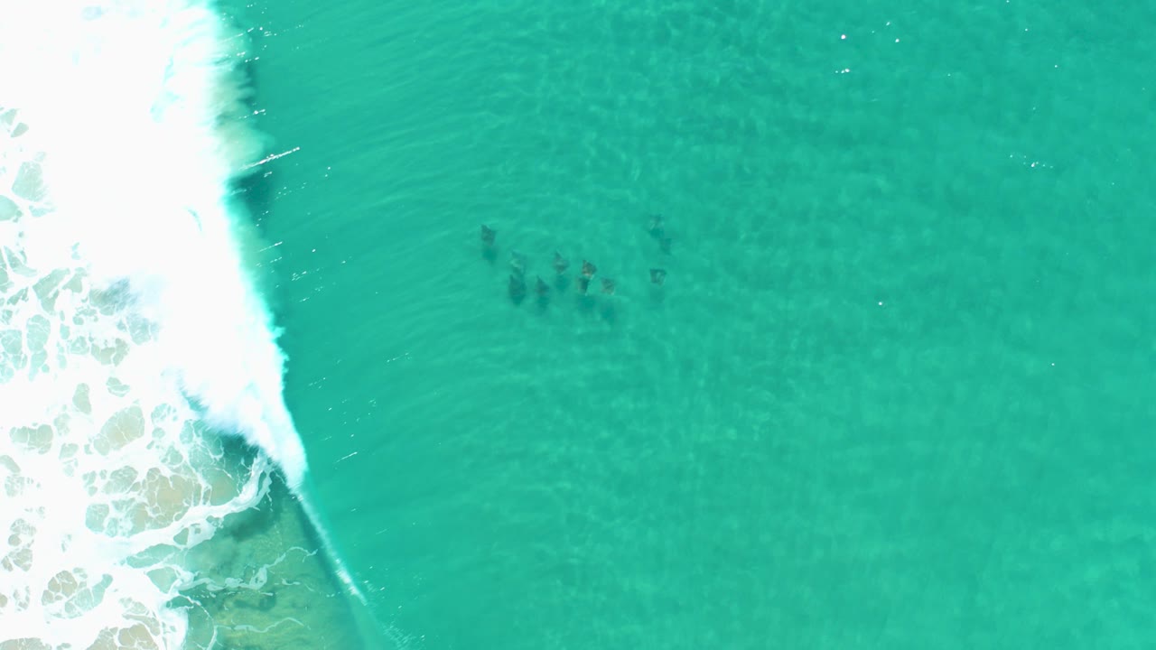 Overhead view of turquoise water in Los Cabos, Mexico, with white surf along the left edge, submerged dark patches near the upper center, and no visible people.