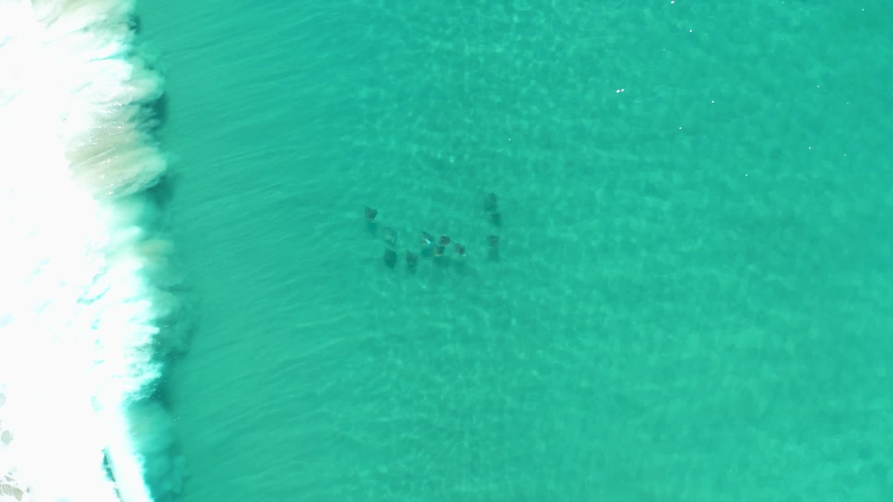 Overhead view in Los Cabos, Mexico, with a brighter band of white wave foam at the left, open turquoise water across most of the frame, faint sand texture, and no visible people.