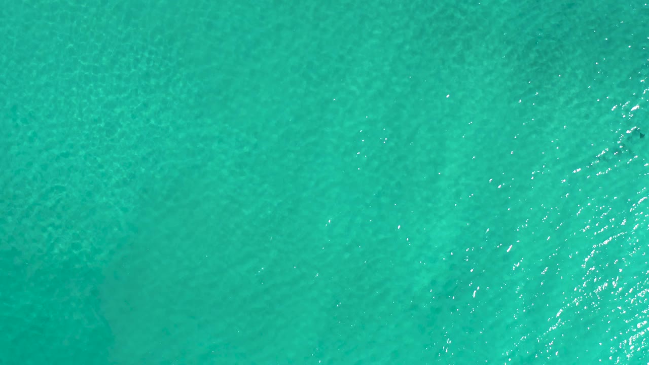 Aerial frame of Los Cabos ocean water with blue-green color, clustered sun reflections near the middle, and no visible people.