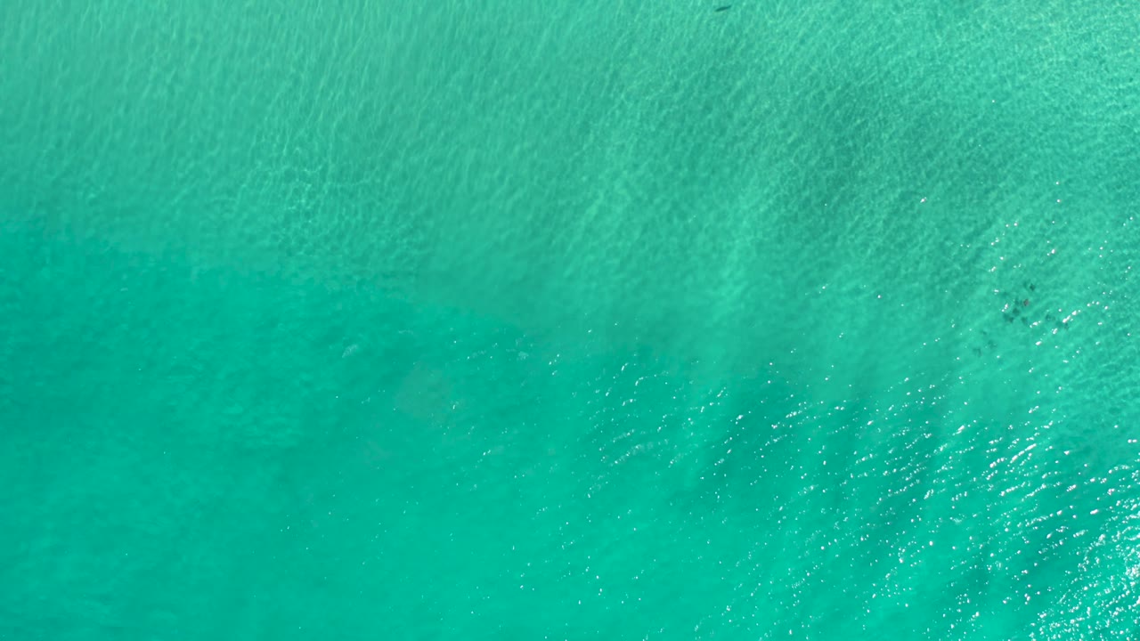 Top-down Los Cabos ocean view showing green-blue water, faint sandy patterns below the surface, small ripples, and no visible people.