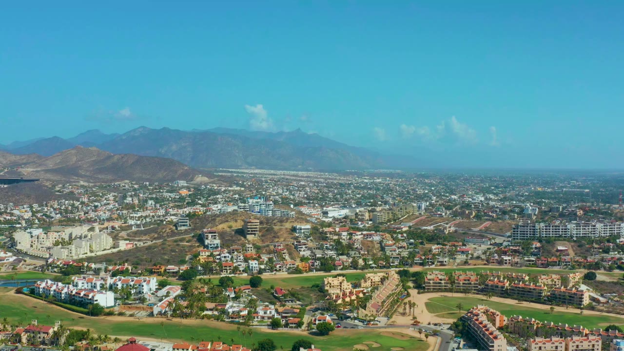 Wide aerial view over Los Cabos, Mexico with white residential buildings, green landscaped areas, dry hills, distant mountains, and no visible people.