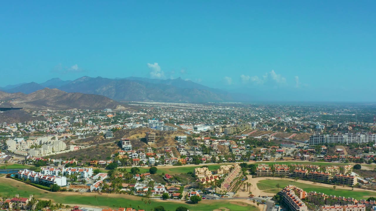 Bright Los Cabos, Mexico aerial panorama with residential blocks, tan hills, green fairway-like strips, and mountains receding into haze, with no visible people.