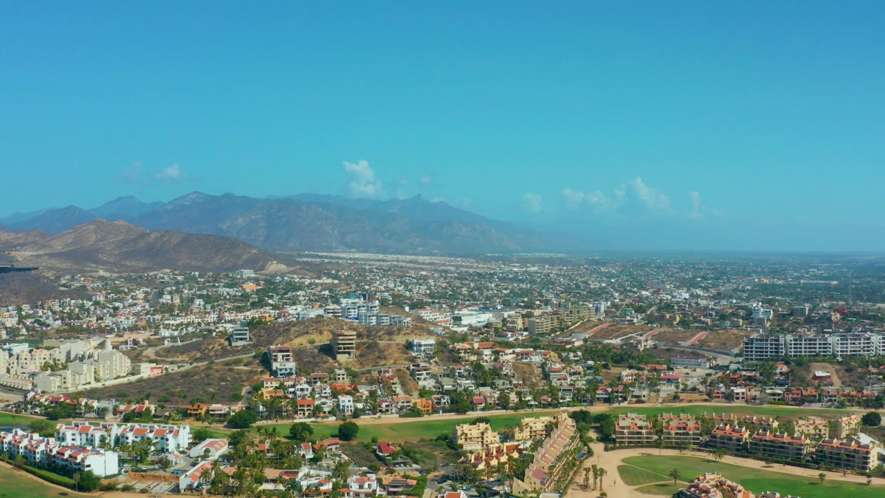 Wide aerial frame over Los Cabos, Mexico, showing hillside houses, low-rise buildings, a green foreground area, dry hills, hazy mountains, blue sky, and no visible people.