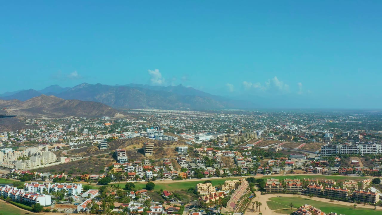 A second wide aerial view of Los Cabos, Mexico, holds on residential blocks, open brown terrain, a green foreground strip, distant mountain ridges, and no visible people.