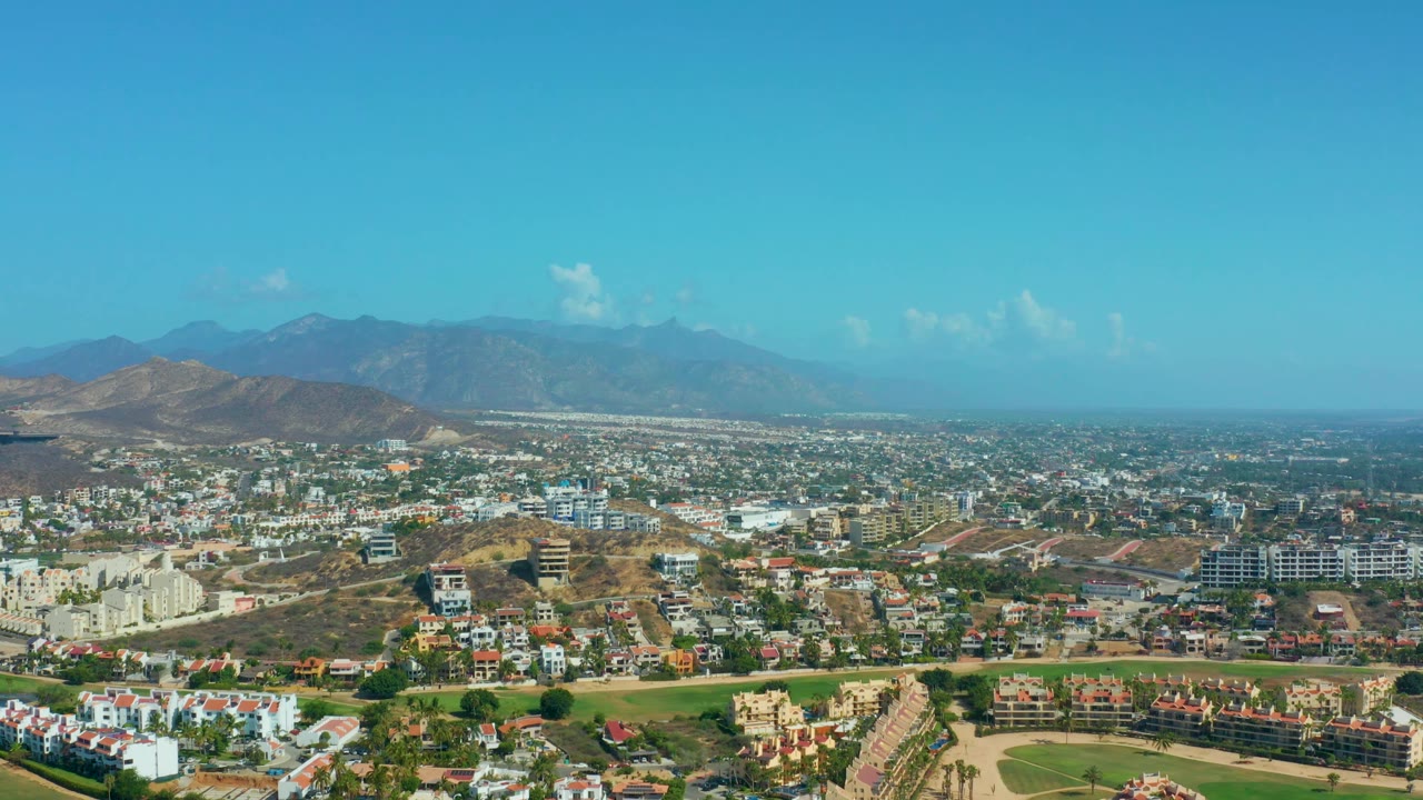 Aerial view in Los Cabos, Mexico, with clustered homes, pale buildings, brown hillside lots, a green foreground area, hazy mountains, and no visible people.