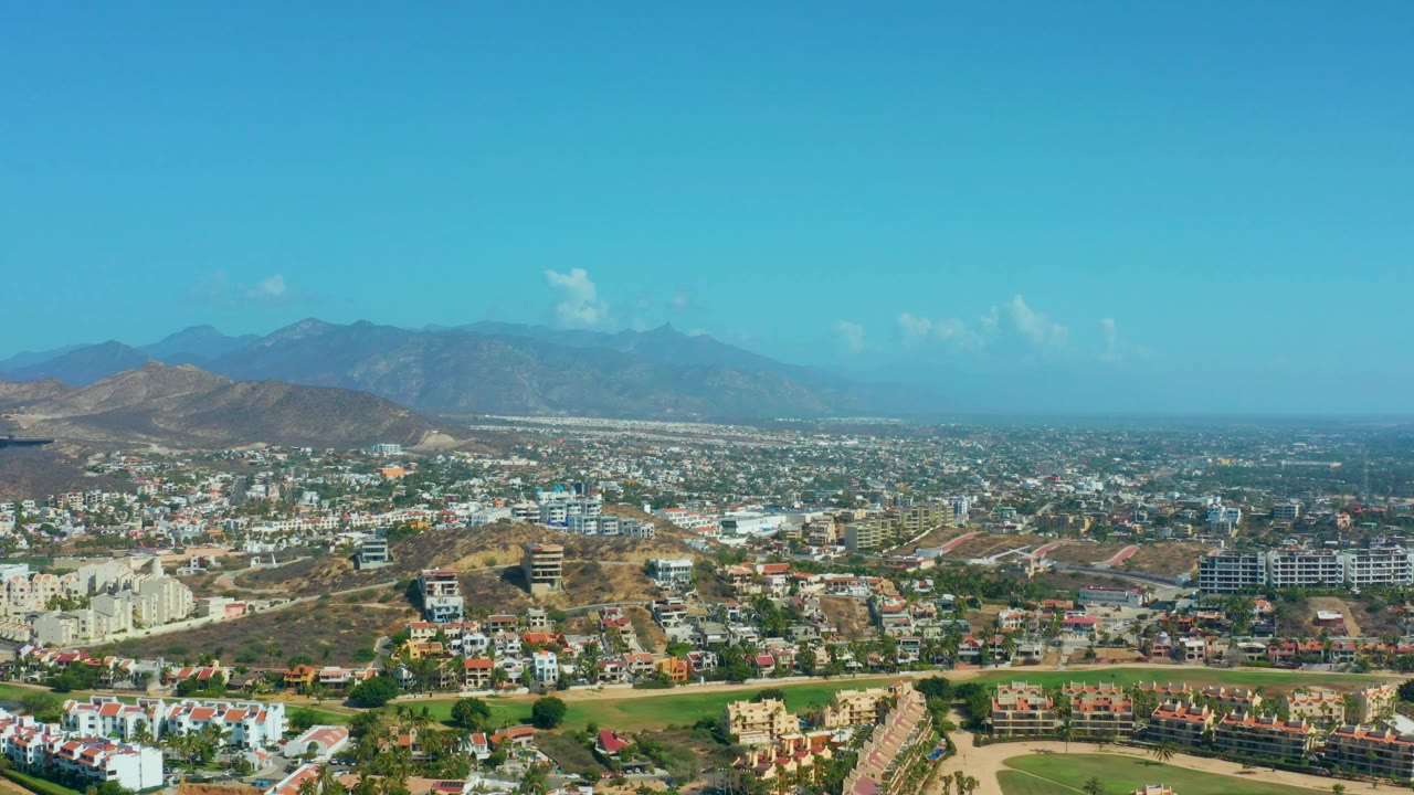 Residential hillside landscape with low buildings, roads, dry slopes, and mountains.