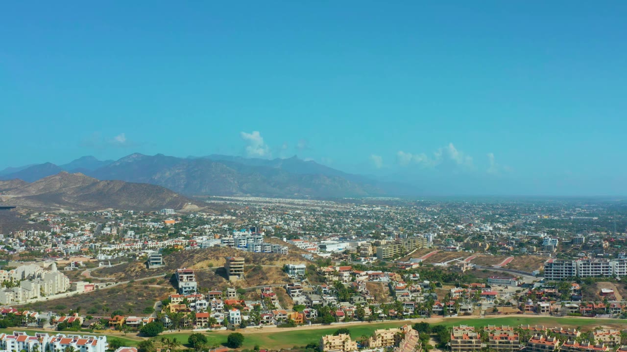 Second aerial frame of Los Cabos showing clustered homes, pale roads, dry hillside lots, and hazy mountains under clear blue sky, with no visible people.