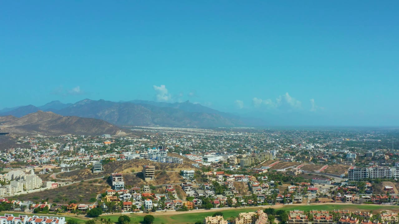 A wide Los Cabos aerial frame with white and tan residential buildings in the foreground, a spread-out town in the distance, and no visible people.