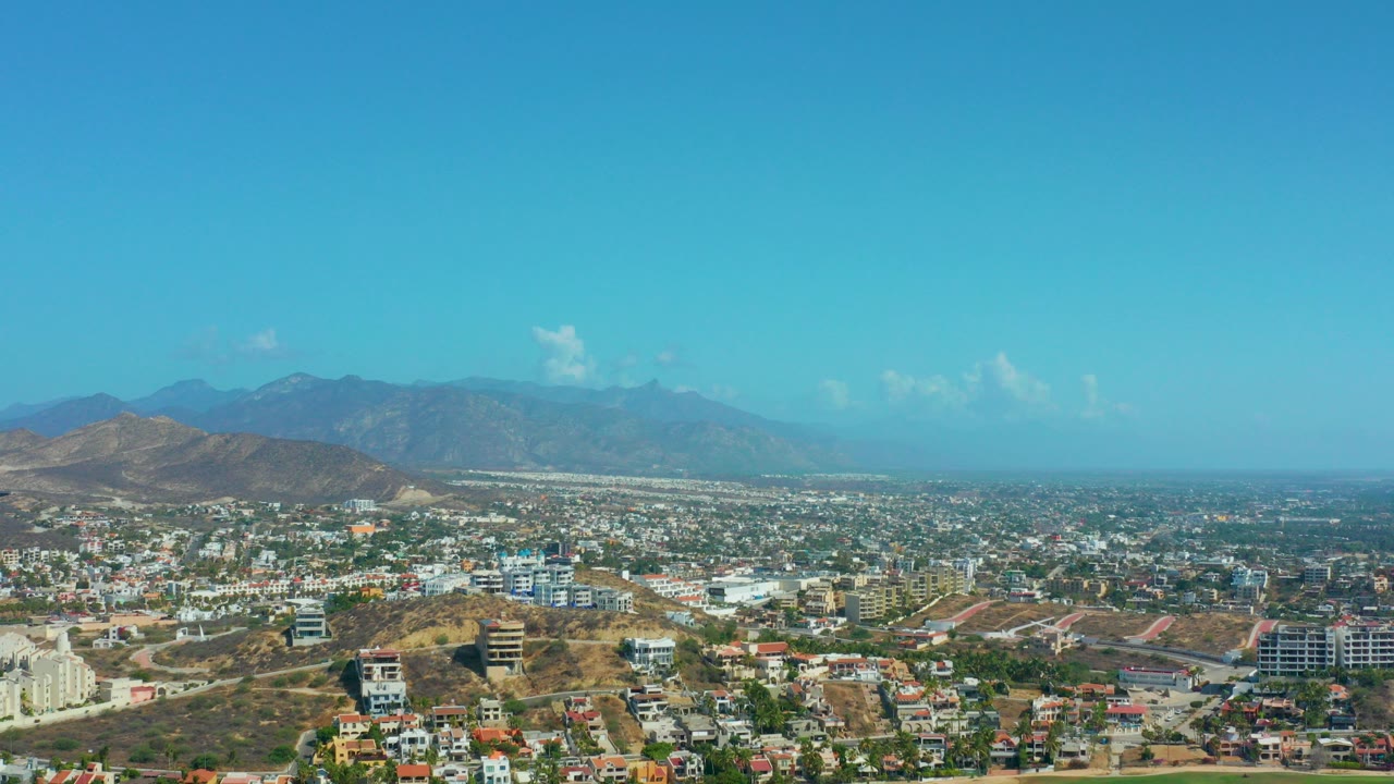 Los Cabos, Mexico aerial view with scrub-covered hills in front, dense city buildings across the middle, distant mountains, scattered clouds, and no visible people.