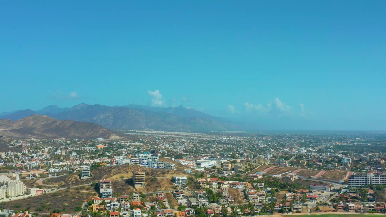 Aerial cityscape with hillside foreground, mountain backdrop, and clear blue sky.