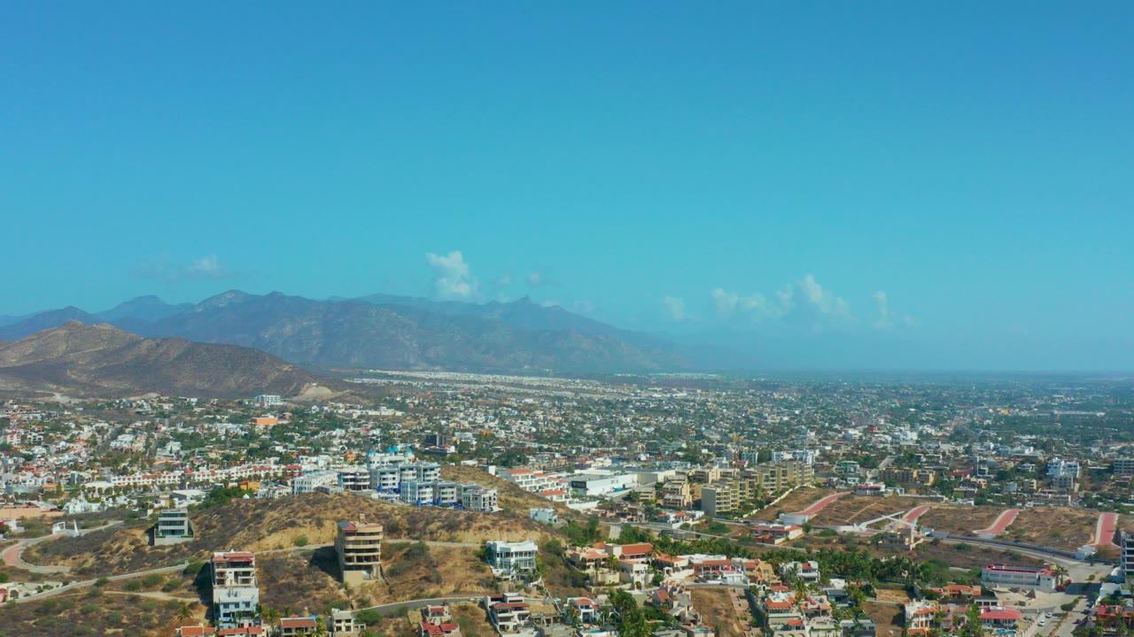 Opening aerial frame over Los Cabos, Mexico with dry foreground hills, low-rise city development, distant mountains, blue sky, and no visible people.