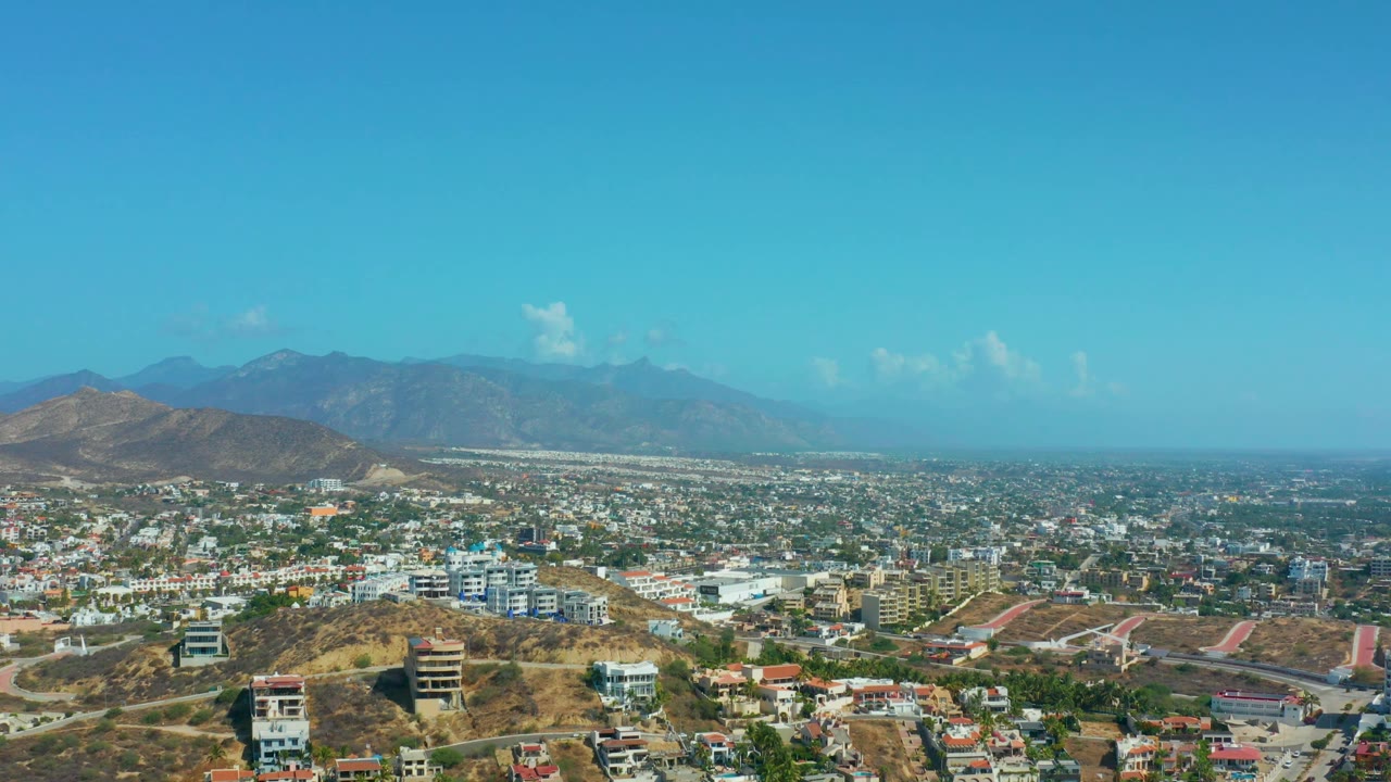 Aerial Los Cabos frame with a broad band of sky above hazy mountains, low-rise buildings across the valley, dry slopes in front, and no visible people.
