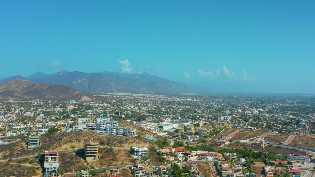 Aerial view over Los Cabos, Mexico with hillside homes in the foreground, dense city buildings in the midground, distant mountains, and no visible people.