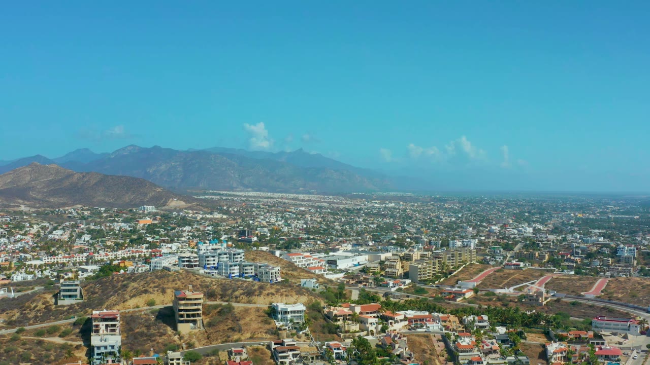 A slightly later aerial frame shows Los Cabos buildings spread across the valley, dry slopes in front, hazy mountains behind, and no visible people.