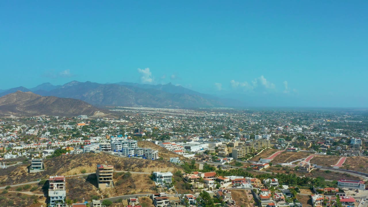 Los Cabos appears from above with clustered white buildings, curving roads, tan hills in the lower frame, a blue sky, and no visible people.