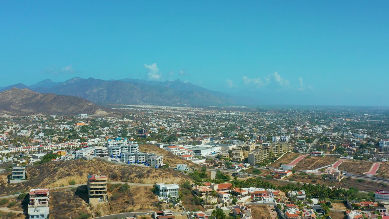Opening frame of Los Cabos, Mexico showing a broad residential cityscape, dry foreground hills, apartment buildings, distant mountains, clear blue sky, and no visible people.