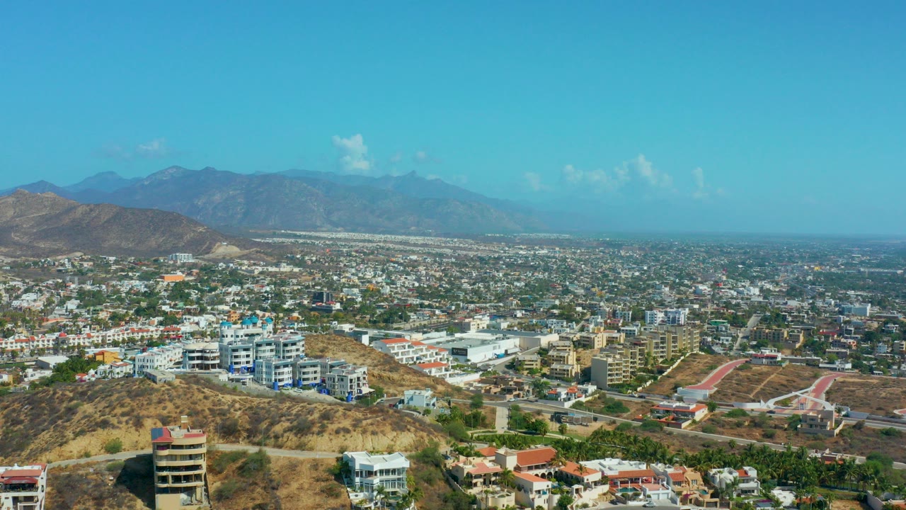 Wide aerial view over Los Cabos, Mexico, with dry foreground hills, scattered buildings, distant mountains, and no visible people at the opening frame.