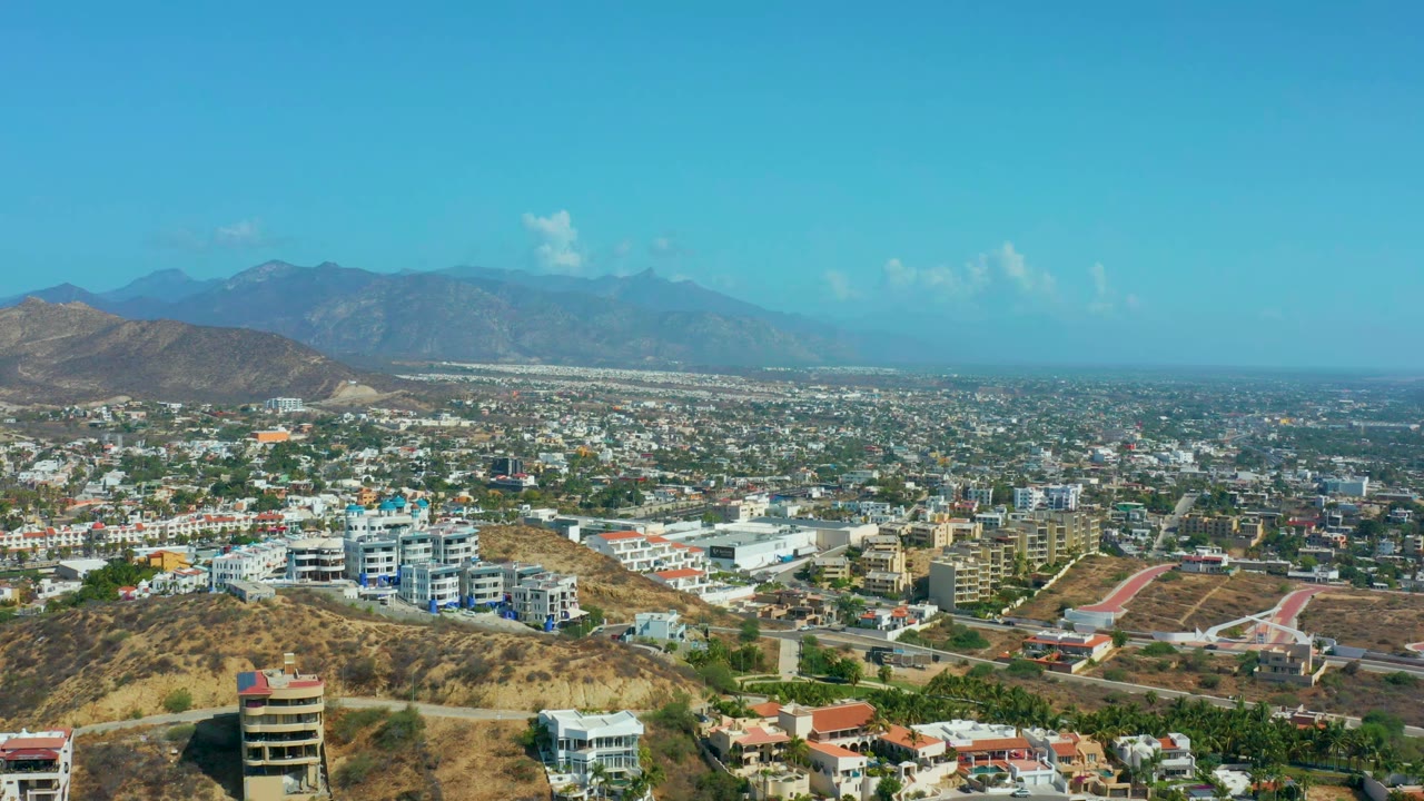 Elevated city view with tan hills, white buildings, roads, and a hazy mountain range.